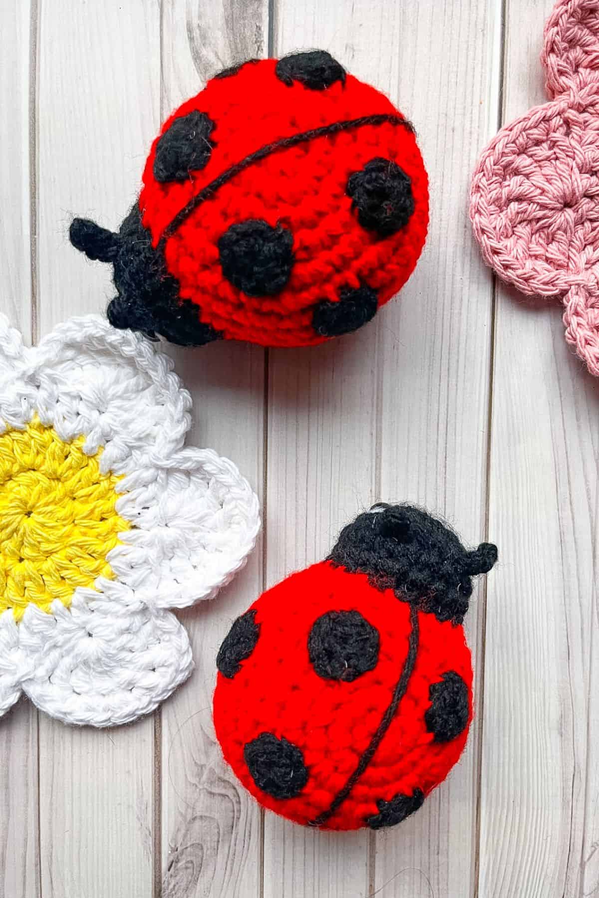 2 finished crochet ladybugs on table with crochet flowers.