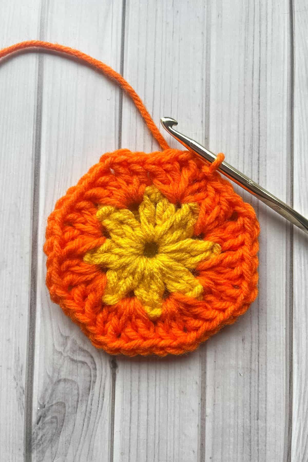 Finished second round of flower granny square, with hook on table.