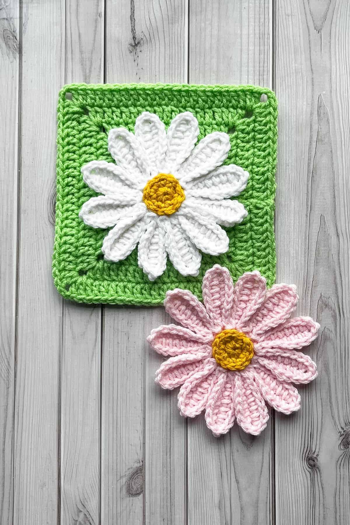 Finished white crochet daisy applique on green square on table, pink daisy is next to it.