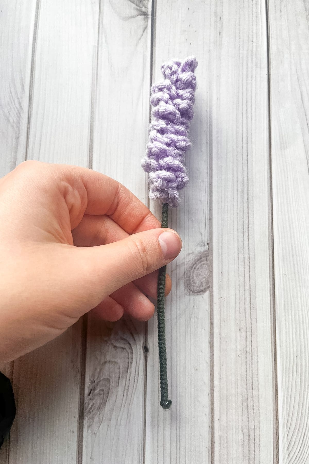 Finished crochet lavender stem in hand above wooden table.